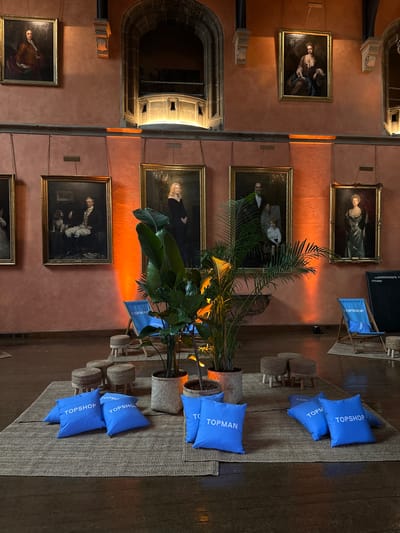 The image depicts an indoor event space with an old-world aesthetic, possibly within a historic building in Chichester, United Kingdom. The walls are a warm, peachy hue, adorned with several large, ornate, framed oil paintings of people in period attire. These paintings are hung at various heights, with some featuring individual portraits and others depicting groups. Below the paintings, a warm orange light emanates from floor-mounted uplighters, casting a dramatic glow on the walls.
In the foreground, a large, textured jute rug is laid out on a dark wooden floor. Scattered across the rug are several bright blue cushions, each emblazoned with white "TOPSHOP" branding. To the right of the cushions, two blue deck chairs with "TOPSHOP" branding are set up. A few woven, round stools are also visible, placed around the rug.
Dominating the center of the scene are two large potted plants with broad, tropical-looking leaves, adding a touch of greenery to the otherwise opulent setting. One plant is in a woven basket planter, and the other is in a terracotta-colored pot.
The overall impression is a blend of historical grandeur and modern branding, suggesting a curated event or exhibition. The lack of people suggests the image was taken before or after the event's main activity.