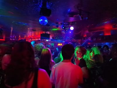 This image depicts a crowded nightclub interior located in Philadelphia, United States. The scene is characterized by dim ambient light intensely augmented by a vibrant array of artificial illumination, primarily featuring blue, red, and green tones. A large, reflective disco ball, prominently lit in blue, is suspended from the ceiling, casting speckled patterns of light. Multiple laser beams, predominantly green and red, traverse the space horizontally through the visible air. Numerous individuals, generally appearing as young adults, are densely packed within the club. In the immediate foreground on the left, a person with dark, blurry hair is partially visible. Centrally, another person wearing a white t-shirt is seen from behind, facing away from the viewer. Toward the center-right, a woman with light-colored, bobbed hair is smiling, her face strongly illuminated by green light, seemingly engaged in conversation or observing someone to her left. Adjacent to her, another woman with long, lighter hair is also bathed in green illumination. The crowd exhibits signs of standing, socializing, and general movement within the energetic club environment. Overhead, various light fixtures and ceiling-mounted equipment are visible.