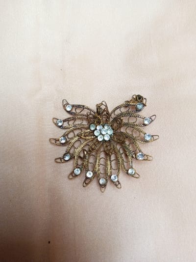 The scene depicts a single antique-style brooch resting on a light peach-colored fabric surface in Qarabucaq qəsəbə, Azerbaijan. The brooch is constructed from aged brass or bronze-toned metal, featuring an intricate filigree design resembling a starburst or stylized flower. Its central element consists of approximately seven small, clear, round faceted stones arranged in a cluster. Radiating outwards from this center are twelve petal-like segments, each formed by openwork filigree and set with a single larger, clear, round faceted stone. The metal exhibits an uneven patina consistent with age and tarnish. A clasp mechanism is visible on the upper right side of the brooch. The fabric background is lightly textured with subtle folds and a slight sheen. No individuals or active interactions are present in the image.