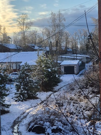 The image captures a static, elevated view of a snow-covered residential or semi-rural area in Kirzhach, Russia, under a partially cloudy winter sky. The foreground exhibits a snow-covered ground interspersed with exposed earth and a winding, partially melted or muddy path extending diagonally from the lower-left. Sparse, bare tree branches occupy the extreme lower-right. Several small, snow-dusted evergreen trees are present in the foreground and midground. In the midground, a light-colored brick garage with a dark, possibly corrugated metal, roll-up door is visible, its roof covered in snow. Behind and to its left, several other single-story structures, consistent with garages or utility buildings, feature snow-covered roofs. A light-colored vehicle, likely a sedan or small SUV, is partially visible parked on a snow-covered surface near a larger building with a white, corrugated roof also bearing a layer of snow. The background reveals additional residential houses with snow-covered roofs, situated amidst numerous bare deciduous trees, which are also lightly dusted with snow. Overhead utility poles and multiple power lines crisscross the upper midground and background. The sky transitions from a bright, hazy yellowish-white on the left, suggesting a low sun angle, to a clearer blue with scattered white clouds on the right. No individuals or dynamic actions are visible.