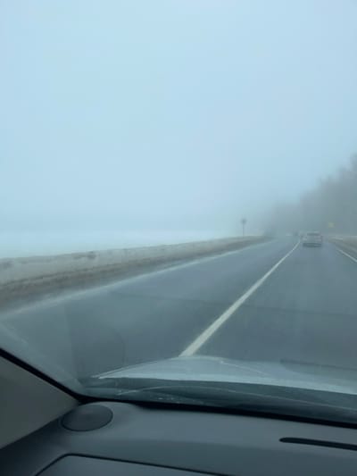 The image depicts a foggy road scene from the perspective of a vehicle's interior, looking forward through the windshield. The lower portion of the vehicle's dashboard and the base of the windshield are visible at the bottom of the frame. The setting is identified as Butyrki, Russia.
The environment is characterized by dense fog, significantly reducing visibility. The sky and distant horizon are a uniform pale blue-grey. The road is a dark, wet or damp asphalt surface, featuring a solid white lane line on its left side, delineating the current lane of travel. To the immediate left of the road, a low, dirty bank of snow or slush runs parallel to the asphalt.
On the right side of the road, a faint outline of trees or vegetation is visible through the fog further in the distance. A circular road sign on a pole stands on the right shoulder, partially obscured by the fog. Multiple vehicles are observed ahead on the road, their forms indistinct due to the atmospheric conditions and distance. At least one vehicle is discernible in the same lane, with additional vehicles further ahead. No individuals or specific actions are identifiable within the scene, which conveys a sense of reduced visibility driving conditions.