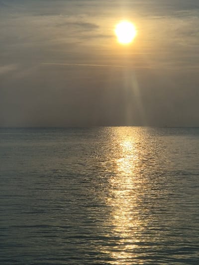 A vertical composition displays a bright sun in the left quadrant, radiating light beams across a misty sky. The right side of the image depicts water, possibly an ocean or lake, reflecting the sunlight in shimmering streaks. The water's surface has small ripples, creating a textured appearance. The overall tone is serene, with a focus on the interplay of light and water.