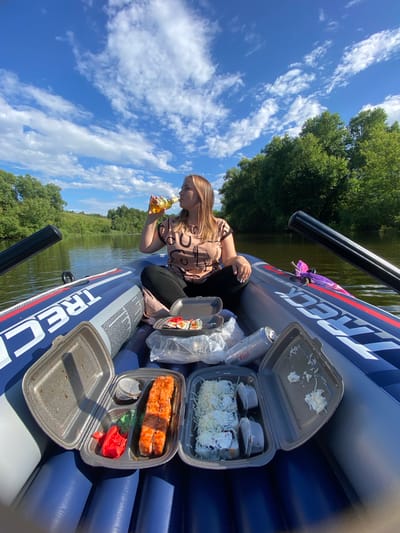 The media file shows a woman sitting in a blue inflatable boat on a body of water, likely a lake or river, given the surrounding trees and vegetation. The sky is a vibrant blue with scattered white clouds, suggesting a clear and sunny day. The woman is wearing a patterned shirt and black pants, and she is holding a bottle of beverage to her lips. In the boat with her are several takeout containers of sushi, some of which appear to be partially eaten. There is also a can of beer and some plastic cutlery. The boat has the word "TRECK" visible in large white letters with a red outline on its side. The overall scene suggests a leisurely outdoor activity, possibly a picnic or a relaxed outing on the water. The time of day appears to be daytime, given the bright sunlight. The location context provided is Chulki-Sokolovo, Russia.