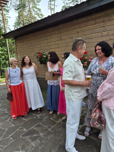 In Zelyonovka, Russia, a group of people are gathered outdoors, standing on a stone-paved walkway in front of a wooden building. The building has a dark-brown, sloped roof and light-brown wooden siding. Red and pink flowers are planted in a dark container on the wall.
On the left, a woman with blonde hair and glasses wears a striped sleeveless top and a long, flowing orange skirt. She holds a small patterned bag in her left hand. Next to her, a woman with brown hair and glasses wears a long, loose white dress.
In the center, a man in a light-colored short-sleeved shirt and white pants holds a glass of beer. He is facing a woman with dark hair, who wears a floral-patterned dress and holds a champagne flute.
Further to the right, another woman in a pink outfit is partially visible. The people appear to be engaged in conversation, suggesting a social gathering or event. The overall atmosphere is casual and relaxed, set against a backdrop of trees.