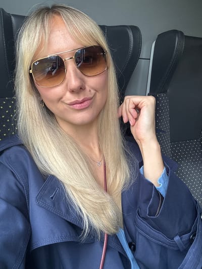 A woman with blonde hair and sunglasses is sitting in a train. She is wearing a dark blue trench coat and a light blue top. She is holding her hand to her face and looking at the camera. The train seats are black with small yellow polka dots. The location is not in Garupe, Latvia.