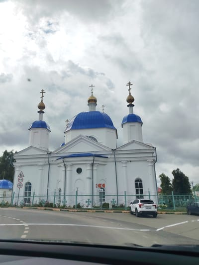 The image depicts a white Orthodox church with blue domes and golden crosses. The church features white pillars and arched windows. In front of the church, there is a light green metal fence adorned with cross-shaped decorations and some rose bushes. A white SUV is parked on the asphalt road in front of the church, facing away from the camera, with its license plate visible. Traffic signs, including one indicating a speed limit of 40, are present near the fence. The sky is overcast with dense clouds. The foreground shows a blurred view of a car's interior, suggesting the photo was taken from a vehicle. The scene is set in the city of Ardatov, Russia.