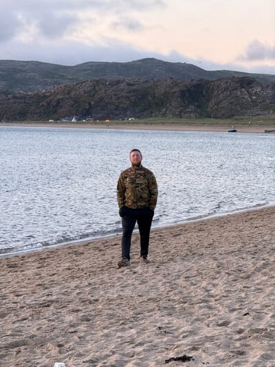 A man stands on a sandy beach in Teriberka, Russia, facing the camera. He is wearing a camouflage jacket and black pants. The beach is next to a body of water, and in the background, there are hills and a cloudy sky. There are several tents and some boats visible along the coastline in the distance. The man has his hands in his pockets and a beard. The sand is textured with ripples and some dark seaweed. The water has small waves.