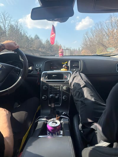 The scene is set inside a vehicle, identified as being in the city of Dobrinovo, Bulgaria, viewed from a front-passenger perspective looking forward through the windshield. Two individuals are partially visible. The driver's right arm, adorned with tattoos on the forearm, rests on a black Volvo steering wheel. The front passenger's legs, clad in dark cargo-style pants, are extended.
The vehicle's interior features a dark dashboard. A red, leaf-shaped "Areon" brand air freshener hangs from the rearview mirror. A pink figurine, possibly a character, is positioned on the dashboard directly in front of the passenger, alongside a black and yellow duck figurine wearing a propeller helmet closer to the center console. The center console includes an infotainment screen displaying "14:40" and Cyrillic text, climate control dials, and an automatic gear shifter. A crushed, open purple Fanta can is situated in a cup holder next to several brown stick-like objects.
Outside the vehicle, an asphalt road is visible, flanked by wooded areas containing bare deciduous trees and some coniferous trees, suggesting a late winter or early spring environment. The sky is bright blue with scattered white clouds. A white truck trailer is observable in the distance ahead on the road, accompanied by a circular road sign on the right side of the road. The overall conditions indicate daytime and sunny weather.