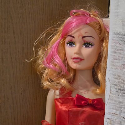 A doll with blonde and pink hair is positioned in front of a wooden background, partially obscured by a white lace curtain. The doll is wearing a red dress with a bow. The doll's face has some marks, appearing to be smudges or dirt. The image does not contain any information about the city of Ravnogor, Bulgaria.