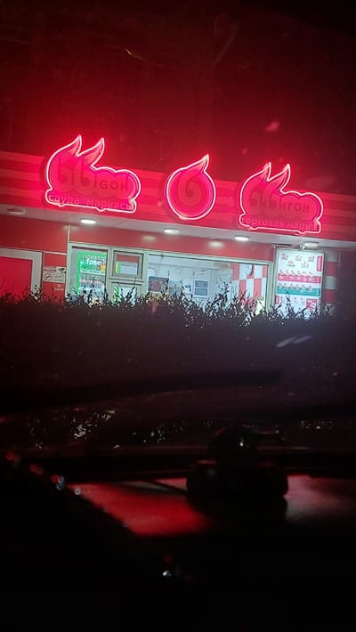 The media file is a photograph of the exterior of a Bibigon store at night. The store has a red facade with bright red neon signs. The neon signs display the Bibigon logo, which appears to be flame-shaped. Text on the sign reads "Bibigon" and "торговая марка" (trade mark). In the background, there is a menu board with pictures of food items and prices. The store appears to be a fast-food establishment. The image is oriented vertically. The time of day is night, as indicated by the darkness and the illuminated neon signs. The setting is urban, likely a street in Shahtinsk, Kazakhstan, as indicated by the text on the sign. There are no people visible in the image. The overall mood is commercial and somewhat inviting due to the bright lights.