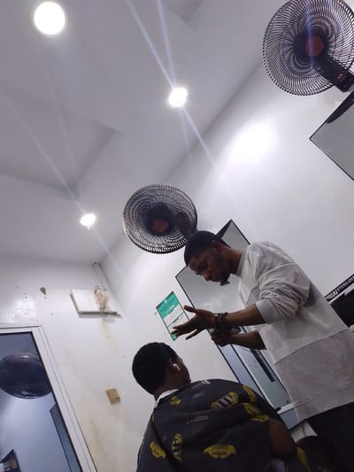 The image depicts the interior of a barbershop, likely located in Abeere, Nigeria, with a focus on a barber attending to a client. The scene is illuminated by overhead circular lights and a ceiling fan is visible in the upper portion of the frame. The barber, a man with dark skin and a beard, is wearing a grey long-sleeved shirt and is in the process of cutting the hair of a client. The client, also a man, is seated and wearing a patterned barber cape. Another ceiling fan is visible on the left side of the image, near a glass-paneled door. The walls appear to be white and there are some posters or signs on the wall, one of which is a green slip of paper with text that includes "PAY WITH TRANSFER" and a phone number. A mirror or a reflective surface is also visible behind the barber. The overall atmosphere suggests a typical barbershop setting.