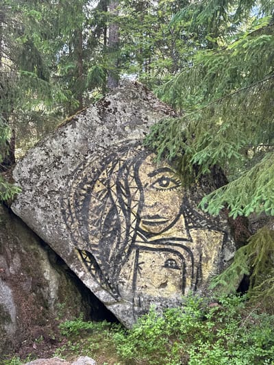 The image shows a large, rough-textured boulder with a striking mural painted on its surface, situated in a forest environment. The mural depicts a stylized face with dark, bold lines outlining features such as large eyes, a nose, and a mouth. The background of the mural appears to be a golden-yellow hue, possibly a base coat applied to the rock. The rock itself is partially covered in moss and lichen, indicating its age and exposure to the elements.

The boulder is nestled among dense coniferous trees, with their branches and needles creating a natural frame around the rock and the artwork. Sunlight filters through the foliage, casting dappled light on the scene. The ground around the boulder is covered with moss, low-lying green plants, and scattered pine needles. The overall impression is one of a natural setting with a man-made artistic element.

The provided context specifies the location as Berezovo, Russia. While the image itself doesn't offer specific markers for a city, the description suggests a rural or forested area within or near Berezovo. There are no people visible in the image. The focus is entirely on the rock and its painted artwork.