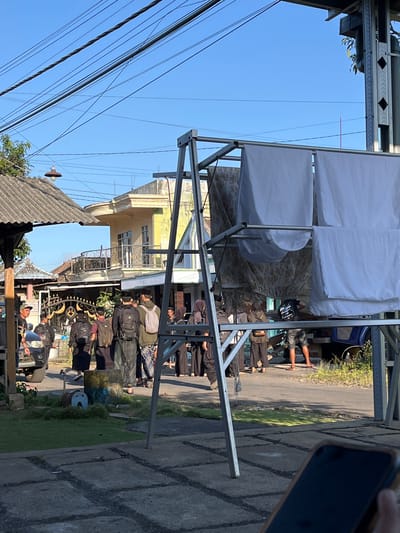 In the city of Jambearum, Indonesia, a group of individuals, most wearing dark clothing and head coverings, are gathered outdoors on a paved area adjacent to a street. The scene is illuminated by sunlight, indicating it is daytime. A collection of overhead power lines crisscross the bright blue sky. In the foreground, a metal A-frame structure holds several white fabric sheets, possibly being used for drying or display, along with patterned fabric panels behind them. A dark-colored car is parked to the left, and behind the group, buildings with balconies and decorative railings are visible. A small, blue and yellow cylindrical object with a face painted on it sits on the grass to the left of the group. The ground is a mix of concrete paving slabs with visible lines and patches of grass. The presence of a mobile phone in the bottom right corner suggests someone is documenting the scene.