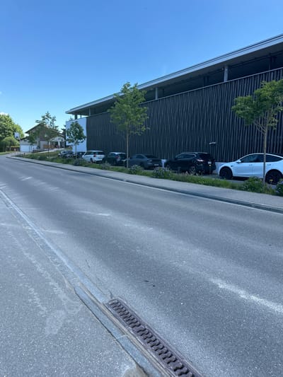 The image depicts a street scene in the city of Le Brassus, Switzerland, under a clear blue sky. The foreground shows an asphalt road with a gutter on the left and a drain cover. Along the right side of the road, there is a row of parked cars, including a white Tesla, a dark SUV, and other dark-colored vehicles. A modern building with a dark, textured facade and a long, overhanging roof is visible behind the parked cars. Trees and landscaping with small purple flowers line the sidewalk in front of the building. Further in the background, on the left, a residential house with a pitched roof and windows can be seen. The scene appears to be captured during the daytime, with no people visible.