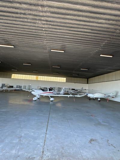 The image displays an interior view of a hangar at an aerodrome, with the provided context indicating Toussus-le-Noble, France. The scene is dominated by several small, white light aircraft parked on a concrete floor.

In the foreground, a white aircraft with prominent red stripe accents is visible, oriented with its nose pointing towards the right side of the frame. Its registration, "F-JDUD," is clearly visible on the tail. Behind it, another similar white aircraft with red details is parked, its tail bearing the registration "F-JAPD." A third, smaller aircraft is partially visible further back, near the entrance.

The left side of the image features a corrugated metal wall, which appears to be dark gray or charcoal. Several rectangular, white light fixtures are evenly spaced along this wall. Higher up on the wall, a bright horizontal strip of light suggests a window or an opening, allowing natural light into the hangar. The floor is made of concrete, showing various scuff marks, stains, and faint lines, indicative of heavy use.

The overall activity depicted is the static storage or parking of aircraft within a hangar. The presence of multiple aircraft and their French registrations "F-JDUD" and "F-JAPD" are key location cues, confirming an aerodrome in France. The lighting suggests it is daytime, with both artificial and natural light sources illuminating the space. No people are visible, and no specific event or activity is actively taking place beyond the stationary presence of the planes.