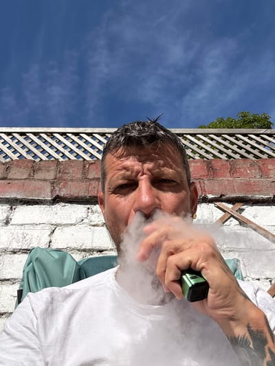 A close-up, over-the-shoulder, selfie-style photograph shows a man exhaling a cloud of vapor from a green electronic cigarette. He is outdoors, sitting in a chair, with a white brick wall and a wooden lattice fence in the background. The sky is blue with some wispy clouds. The man has short, dark, spiky hair and is wearing a white t-shirt. He has tattoos on his arm and is wearing a gold earring. The image is well-lit, suggesting it was taken during the daytime. The location is identified as Warrington, United Kingdom.