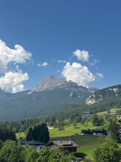 The image depicts a scenic mountain landscape in Cortina d'Ampezzo, Italy. The scene is dominated by a majestic, rocky mountain peak with a dense forest covering its lower slopes. The mountain face is rugged and imposing, with visible rock formations and crevices. A clear blue sky with scattered white cumulus clouds stretches above, suggesting fair weather and a bright, sunny day.
In the foreground and mid-ground, a well-maintained green lawn slopes downwards, dotted with several modern buildings, appearing to be houses or chalets. These structures have dark roofs and visible windows. The architecture suggests a residential or resort area nestled within the natural beauty of the mountains. The presence of the manicured lawn and buildings indicates human habitation and development in this mountainous region.
The sunlight casts distinct shadows, indicating it is likely midday or early afternoon. The overall atmosphere conveyed is one of tranquility and natural grandeur, with the human presence being integrated harmoniously into the stunning alpine environment. There is no visible text in the image.