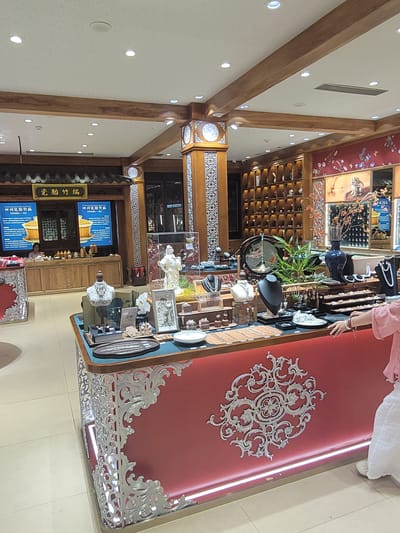 This image shows the interior of a jewelry store, likely located in Wuhou District, China, as suggested by the signs with Chinese characters. The store has a traditional Chinese aesthetic with wooden beams, intricate carvings, and decorative artwork on the walls. The main display area features a long counter with various jewelry items, including necklaces, bracelets, and pendants, presented on busts and trays. A person, possibly a customer, is partially visible on the right side of the frame, interacting with the display. In the background, shelves are lined with more decorative items, and screens display information about the products. The lighting is warm and inviting, highlighting the merchandise.