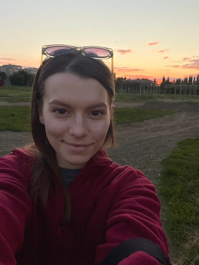 A young woman with dark hair and sunglasses perched on her head takes a selfie outdoors at sunset. She is wearing a burgundy fleece jacket over a dark shirt. The background features a dusty, gravelly area with patches of green grass. In the distance, there are some buildings, trees, and a skeletal structure that appears to be under construction. The sky is a gradient of warm colors, from pale yellow to orange and pink, indicative of a sunset. The image was taken in Kotovo, Russia.
