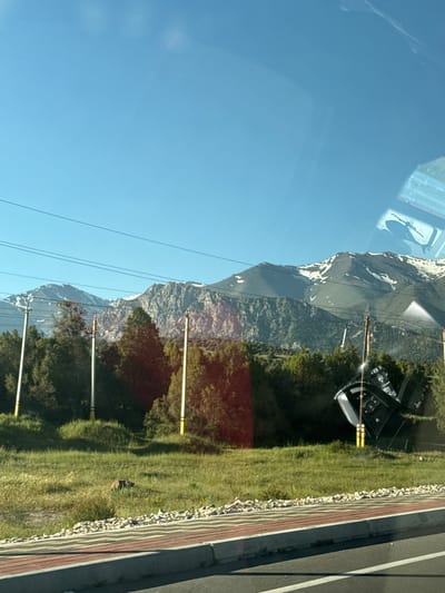 The image shows a landscape scene with mountains in the background and trees in the foreground. There are power lines running across the sky. In the middle ground, there are several utility poles. To the right, a black car is overturned on its side, resting against one of the utility poles. The car appears to be damaged. A road with a brick border is visible in the lower part of the image. The scene is set under a clear blue sky. The provided location "Myk, Uzbekistan" is not verifiable from the visual information in the image.
