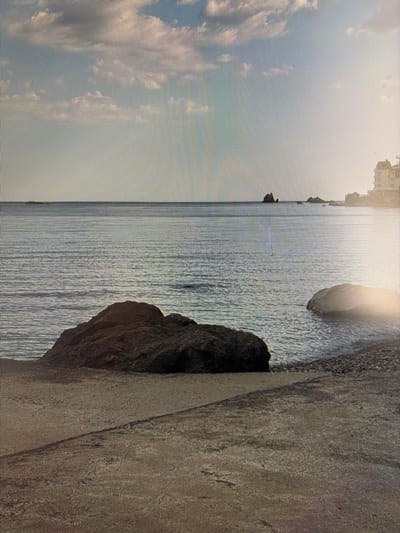 The image depicts a serene coastal scene. In the foreground, a rough, textured concrete walkway or pier leads towards the water. To the left, a large, dark, jagged rock is partially submerged at the water's edge. Further out in the calm, shimmering sea, several smaller rocks and islets are visible. On the horizon, a silhouette of buildings can be seen on the right side. The sky is a pale blue, with scattered, soft white clouds catching the gentle light. The overall impression is one of peaceful natural beauty, with hints of human presence on the distant shore. The image does not contain any people or specific identifiable landmarks that would place it in Mirnoye, Ukraine.