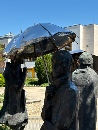 A bronze sculpture in Siófok, Hungary, features three human figures under a deconstructed metallic umbrella. The figures, rendered in a dark, rough texture, are positioned closely together. The umbrella, seemingly incomplete with exposed struts, contrasts with the organic forms of the figures. The background includes a building with windows, greenery, and a patterned walkway composed of stone and brick. The sky is clear and blue.