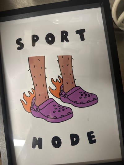 A vertically oriented, black-framed print with a clear cover displays a white background featuring a cartoon illustration. The top of the print reads 'SPORT' in bold, rounded black capital letters. Below this, two light brown cartoon legs with short black hairs are depicted, each wearing a purple, perforated, Crocs-style sandal. Orange flames emanate from behind the heels of both feet. The bottom of the print reads 'MODE' in matching bold, rounded black capital letters. The framed print is positioned against an indistinct background surface, with a segment of a white coiled cable visible in the upper right. This scene is located in the city of Salisbury, United Kingdom.