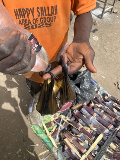 An outdoor, eye-level shot in Rijiyar Lemo, Nigeria, depicts a person's upper torso and arms. The individual wears an orange short-sleeved t-shirt with the black text "HAPPY SALLAH AREA OF GROUP 2025" printed across the chest. Their left hand holds a clear plastic bottle, partially obscuring a red and black label, tilted to pour a transparent liquid into one of two small black plastic bags. These bags are tied at the top with black material and are held by the fingers of the person's right hand. Each bag contains a visible block of yellowish solid food. To the lower right, a large quantity of cut sugarcane stalks, exhibiting reddish-purple and white sections, is piled on a surface, partially covered by green netting. A red bowl is partially visible near the base of the black bags, resting on the sugarcane. The ground is light brown dirt, illuminated by bright sunlight casting distinct shadows.