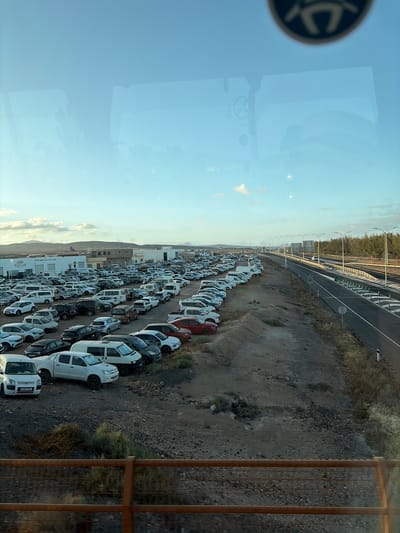 The image shows a large parking lot filled with primarily white vans, located next to a highway. The vans are parked in rows on a dirt surface. In the background are buildings and hills, potentially indicating an industrial or commercial area. The overall scene is shot from an elevated perspective, possibly from a building window, with part of the window frame visible. Given the clues present such as the website on the side of one of the vans, it is likely the image shows the industrial estate of Puerto del Rosario, Spain.