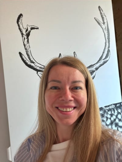 The image shows a selfie of a Caucasian woman with blonde hair, blue eyes, and a wide smile. She is wearing a striped shirt over a white t-shirt. Behind her, on a large white canvas, is a black and white illustration of deer antlers. The lighting suggests it is daytime. There is no visible indication of the specific location within the city of Sauris/Zahre, Italy.