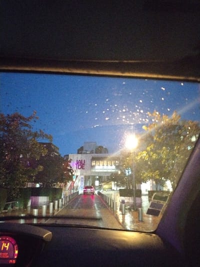 The image presents an urban evening scene viewed from the interior of a vehicle, looking forward through a rain-splattered windshield. The vehicle's dashboard, visible in the lower foreground, features a digital display showing "OCTOBRE" and "19:15" (or "19:16"), indicating the time and month. The central focus is a multi-story building displaying the prominent, illuminated sign "JAUDE" in large letters, identifying it as Centre Jaude in Clermont-Ferrand, France. A wet, reflective paved path, bordered by numerous silver cylindrical bollards, leads directly towards the building's entrance. A dark-colored sedan with illuminated red taillights is positioned centrally on this path, facing away from the viewer. The environment is illuminated by streetlights, which cast reflections on the wet ground, and the building itself is brightly lit. Trees with a mix of green and autumnal yellow foliage are visible on either side of the path. The sky is a deep blue, characteristic of dusk or night. No people are visible.