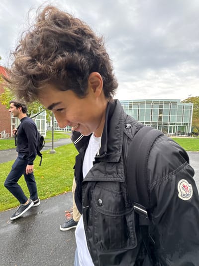 The image captures two males walking on a paved pathway at Wellesley College in Wellesley, Massachusetts. The Lulu Chow Wang Campus Center, a multi-story, curved glass and steel building, is visible in the mid-ground, with a traditional brick building partially visible on the far left. The sky is overcast.

In the foreground, a young male with voluminous dark curly hair is seen in profile, smiling and looking downwards. He wears a black Moncler jacket with the brand's logo patch on the left sleeve, a white t-shirt underneath, and beige trousers. A black backpack strap is visible over his right shoulder.

Behind and to his left, a second male walks on the same path, carrying a black backpack. He wears a dark long-sleeved top, dark trousers, and dark slip-on shoes with white soles. He is also smiling and looking forward.

The paved path appears wet, and green grass flanks the walkway. Scattered autumn leaves are present on the ground.