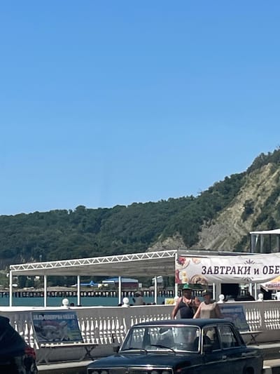 The image depicts a sunny day scene with a clear blue sky. In the foreground, a dark-colored vintage sedan is parked. Behind it, a white fence with decorative elements lines a promenade or walkway. Two people, a man and a woman, are visible walking near a banner that reads "ЗАВТРАКИ И ОБЕ" (Breakfast and Lunch). To the left of the banner, a table displays a poster with an image of animals. Further back, a covered structure with a white, triangular roofline extends over an area with tables and chairs, suggesting an outdoor dining or seating area. In the background, a densely wooded hillside rises steeply, and beyond that, a body of water can be seen, likely the Black Sea, with structures and a pier extending into it. The environment appears to be a coastal resort area. This scene is consistent with locations found in Lermontovo, Russia, a popular seaside resort town on the Black Sea coast.