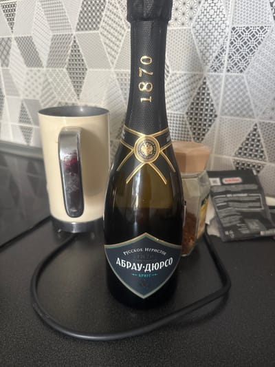 This image shows a bottle of Abrau-Durso Brut sparkling wine placed on a black surface, likely a table. The bottle is dark green and features a gold and blue label with Cyrillic text. The label prominently displays "РУССКОЕ ИГРИСТОЕ" (Russian Sparkling) and "АБРАУ-ДЮРСО" (Abrau-Durso), along with the year "1870". A golden emblem with the Russian coat of arms is also visible on the neck label.
In the background, a patterned surface with geometric designs in grey and white can be seen, resembling a kitchen backsplash or tablecloth. To the left of the bottle, a portion of a beige appliance, possibly a kettle, is visible. Behind the bottle, there are a couple of packets, one of which appears to be a dark cardboard box with some text, and a glass jar containing what looks like coffee beans. The lighting suggests an indoor setting, possibly during the day or under artificial light. No people are present in the image, and the scene appears to be a still life arrangement. The overall impression is that of a casual, domestic setting.