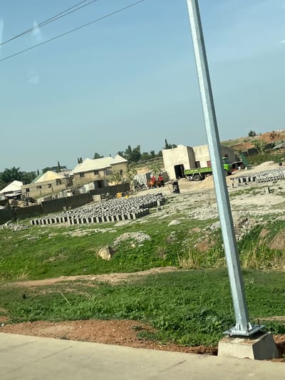 This image captures an outdoor scene at a construction or development site in Uke, Nigeria, under a clear, bright sky. The photograph is taken from a slightly elevated viewpoint, looking down upon the area. A prominent, silver metallic pole, likely a street light or utility pole, runs diagonally from the lower right foreground towards the upper left, partially obscuring the view.
In the midground and background, several unfinished buildings constructed from concrete blocks are visible. Some of these structures have rudimentary roofs, suggesting residential or commercial development. Adjacent to these buildings and spread across the site are large, neat stacks of grey concrete blocks, indicating active construction. Two green trucks are present: one is partially hidden behind a building on the left, and another is visible further back within the site.
The ground is a mix of bare earth, dirt paths, and patches of green grass and weeds. There are also areas with scattered debris and rubble typical of a construction zone. The weather appears sunny with a light blue sky, indicating daytime, possibly late morning or early afternoon, given the lack of harsh shadows. No people are visible within the frame, and there is no discernible text. The scene conveys ongoing development and building activity.