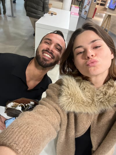 A selfie-style photograph captures two individuals, a male and a female, within an indoor commercial establishment, identified as being in Assevillers, France. The female, positioned in the foreground right, has brown hair and is wearing a light-brown textured jacket with a prominent light-colored faux fur collar. She is making a "kiss" face with pursed lips and holds a smartphone, partially visible in the lower-left, which displays a keyboard interface. To her left and slightly behind, a male subject with dark hair, a beard, and a black and white patterned headband is smiling broadly, revealing his teeth. He wears a black collared shirt.
The background features a bright white counter or table surface. On this surface, a brown pouch containing charging cables and a white adapter is visible near the male. Further along the counter, a small dark disposable coffee cup, a brown paper bag, and a stack of pink boxes are present. A person, seen from the back wearing a dark puffer jacket and light-colored trousers, stands near the counter in the far-left background. Distant wooden-toned shelving and a vertical display poster are also discernible in the background. The flooring appears to be light-colored.