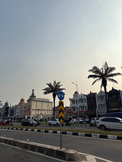 The image captures a street scene in Pakulonan Barat, Indonesia, with a row of multi-story buildings lining the right side of a road. The buildings exhibit diverse architectural styles, including ornate facades with balconies and what appear to be decorative domes and spires. Palm trees are interspersed among the buildings. Several vehicles, including cars and motorcycles, are present on the road, with some in motion. A traffic sign with blue and white symbols is visible on a pole. The sky is overcast, suggesting it could be morning or late afternoon. The foreground shows a paved road with a concrete barrier and grass median. There are no clearly identifiable people in focus, though movement suggests activity. The overall impression is of a developed urban commercial area.