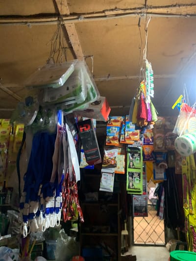 This media file depicts a dimly lit indoor scene, likely a small shop or stall in Farin Yashe, Nigeria. The primary subjects are a variety of packaged goods, including what appear to be tea packets (Lipton, Peak), coffee sachets, snacks (Ginmil), and possibly some electronic accessories like headphones. Colorful decorative items, possibly umbrellas or tassels, are also prominently displayed, hanging from wires. The setting appears to be a storage area or a market stall, with shelves and hanging displays. The lighting is artificial and creates a somewhat cluttered and shadowed atmosphere. There is no discernible activity or event taking place, suggesting a still shot of the merchandise. Notable text visible includes brand names like "Lipton," "Peak," "Ginmil," and "Coffer," as well as product descriptions such as "POWERFUL BASS HEAD" and "TEREO HEADPHONES." The overall impression is one of a bustling but modestly organized local retail environment.