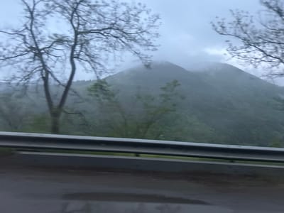 The image shows a mountainous landscape near Lukovo, Bulgaria, viewed from a road. The foreground contains a metal guardrail and a wet road surface, likely due to rain. Beyond the guardrail, there are trees with bare branches and a verdant hillside leading to fog-covered mountains in the background. The sky is overcast, suggesting a cloudy or rainy day. The overall scene is blurred, possibly indicating that the image was taken from a moving vehicle.
