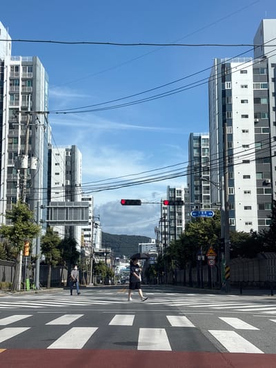 The image depicts a street scene in Cheongju-si, South Korea. The foreground features a crosswalk with distinct white stripes on a dark asphalt road. Two individuals are crossing the street from left to right. The person on the left is wearing a light gray t-shirt, blue jeans, a crossbody bag, and a face mask, and is walking with their hands in their pockets. The person on the right is wearing a black t-shirt, black shorts, and is holding a black umbrella over their head.
In the mid-ground, traffic lights are visible, with the red light illuminated. A street sign with Korean characters and "Juseong-ro" in English is mounted on a pole. Several trees line the street, interspersed with utility poles and overhead power lines. Various buildings, including tall apartment complexes with numerous balconies, are prominent on both sides of the street. The buildings have a modern architectural style, characterized by white and gray facades.
The background shows more urban structures, a hilly landscape, and a clear blue sky with some wispy clouds. The overall environment suggests a daytime scene in a developed urban area.