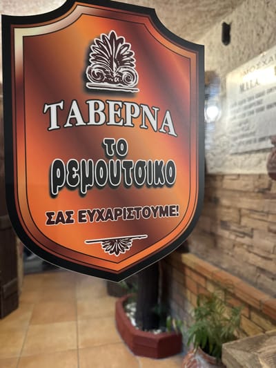 The image displays a decorative, shield-shaped sign for a tavern, set within what appears to be an establishment in Agios Dimitrios, Greece. The sign, oriented vertically, features a warm gradient background transitioning from a rich dark brown at the top to a vibrant orange at the bottom.

Prominently displayed in white, outlined Greek text are the words: "ΤΑΒΕΡΝΑ" (Taverna), followed by "το Ρεμπουτσικο" (to Remboutsiko), which is likely the name of the establishment. At the bottom, a welcoming message reads "ΣΑΣ ΕΥΧΑΡΙΣΤΟΥΜΕ!" (Thank you!). Two stylized white motifs adorn the sign: a more elaborate, classical-looking design resembling an ancient Greek column capital or acanthus leaf on the left, and a simpler, smaller decorative element on the bottom right.

In the background, a wall constructed from light-colored, irregularly shaped stone or brick-like tiles is visible. The floor beneath is a light-toned tile. To the right of the sign, a green potted plant adds a touch of natural decor to the setting. In the upper left background, part of a blurred paper with some unreadable Greek text, including the word "ΜΟΣΧΑ" (Moscha), is faintly visible. The overall lighting suggests an indoor or a shaded outdoor environment.