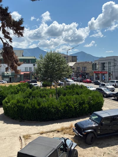 The image shows a street scene in Khashuri, Georgia, under a bright, cloudy sky. In the foreground, a dark-colored Jeep Wrangler is parked, with its roof up. To the left, the roof of another dark-colored Jeep Wrangler is visible. A circular, well-maintained evergreen hedge dominates the middle ground, with a single tree growing within its center. Beyond the hedge, a street is lined with buildings of various architectural styles. Several cars are parked and driving on the street, including a red sedan, a black SUV, and a white van. Mountains form the backdrop of the scene, their peaks softened by distance and the haze of the day. The overall impression is of a typical urban environment in a mountainous region, with a clear day and moderate activity.