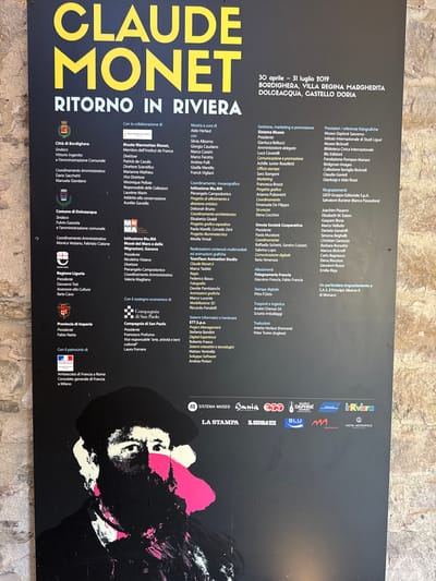 This is a photograph of a large black poster advertising an exhibition of Claude Monet's work titled "Ritorno in Riviera." The exhibition is taking place from April 30 to July 31, 2019, at various locations including Bordighera, Villa Regina Margherita, and Dolceacqua, Castello Doria. The poster features a stylized, graphic representation of Claude Monet's face in black, white, and pink. The text on the poster is primarily in white and yellow, with some smaller sections in other colors. The poster is mounted outdoors, as evidenced by the rough, textured stone wall visible to the right and behind it. The lighting suggests it is daytime. The poster itself is the main subject, with the surrounding environment providing context.