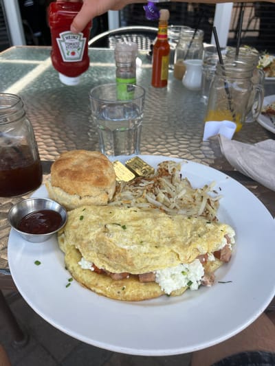 The scene is a close-up, overhead shot of a breakfast plate on a glass-topped table, likely outdoors. A hand is partially visible, holding a bottle of Heinz Ketchup.
The plate contains a large omelet with what appears to be ham and cottage cheese filling, hash browns, a biscuit, butter chips, and a small container of sauce, possibly jam.
Other items on the table include a jar filled with a yellow liquid and straws, presumably juice, a glass of water, a bottle of hot sauce, a salt shaker, and a jar of coffee.
The reflective surface of the table shows the scene is outdoors. Based on the style of food and ambiance of the setting, it is likely taken in Palm Desert, United States of America.