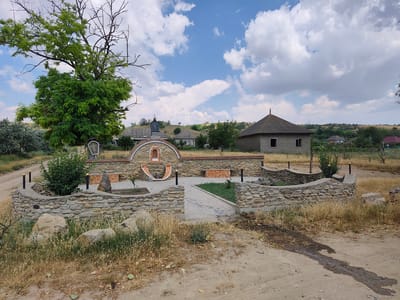 The image depicts an outdoor scene in a rural or village setting, likely in or near Alexandru Ioan Cuza, Moldova. In the foreground, a circular, low stone wall encloses a small courtyard with a paved walkway leading into it. Within the courtyard, there is a decorative stone structure featuring an arched recess with mosaic tiles and a small basin. Benches with mosaic tile surfaces are also visible. A large, leafy tree stands prominently to the left of the courtyard. In the background, several modest houses with pitched roofs are scattered across rolling, dry, grassy hills under a bright blue sky dotted with white clouds. The overall impression is one of a small, somewhat rustic community on a sunny day.