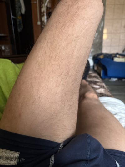 A close-up, eye-level shot focuses on a person's thigh and groin area, covered by navy blue underwear with white stitching. The thigh is covered in light brown hair. The background is blurred, showing a green blanket on the left, a bed with white sheets on the right, and a blue piece of furniture in the distance. The overall scene appears to be indoors, likely in a bedroom. There is no visible indication of the location in L'gov, Russia.