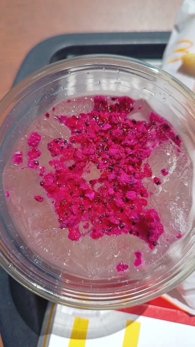 A close-up, overhead shot captures a clear plastic cup filled with ice and a pink beverage. Scattered across the top of the ice are finely diced pieces of pink dragon fruit, interspersed with small black seeds. The cup is placed on a gray surface, possibly a tray, which is partially visible in the background along with a white paper wrapper with yellow and red stripes. The scene is indoors, with soft lighting.
