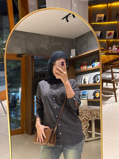 The image displays an indoor scene in Patrang, Indonesia, captured via a mirror selfie. A female individual is reflected in a large, vertically oriented mirror with an arched top and a gold-colored frame. She wears a dark hijab, light-framed glasses, a dark grey long-sleeved button-up shirt with a subtle geometric pattern, and light blue denim jeans. She holds a light pink smartphone in her right hand, obscuring her upper face as she takes the photograph. A brown leather crossbody bag is held in her left hand, and a gold-colored bracelet is visible on her right wrist, with a ring on her left ring finger.

The environment reflected around her includes grey textured walls with warm ambient lighting, suggesting a cafe or restaurant setting. To the left of the reflection, wooden double doors are partially open, revealing an illuminated outdoor area with foliage, indicative of evening conditions. A dark speaker unit stands near the doors. To the right of the reflection, wooden shelves with integrated yellow backlighting display multiple vinyl records featuring various album covers. A white surveillance camera and a small white electronic device are mounted on the wall. In the lower right corner, a wooden table with a patterned tablecloth and a wooden chair with a light grey cushion are visible, along with several containers with red tops on the table. The floor is light-colored tile.