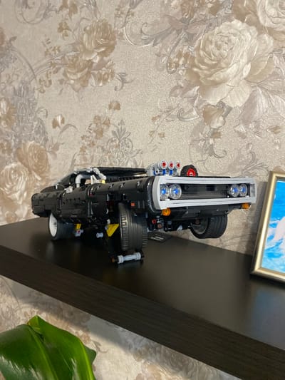 A black Lego replica of a classic car is displayed on a dark wooden shelf against a floral wallpaper background. The car has visible engine details, including four exhaust pipes on top and detailed headlights. A framed picture is partially visible to the right of the car. A lush green plant is in the foreground, partially obscuring the bottom left of the shelf. The setting appears to be an indoor domestic environment.