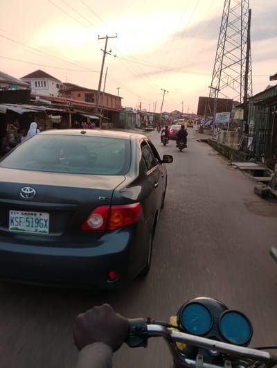 The image depicts a street scene in Isheri, Nigeria, during what appears to be sunset or early evening, indicated by the warm tones of the sky. A dark-colored sedan, possibly a Toyota Corolla based on the badge and taillight design, is prominently featured in the foreground, viewed from the rear. Its license plate reads "KSF-519GX" and is from Lagos. The car is on a paved road lined with buildings and structures on both sides.
To the left of the road, several people are visible near market stalls or small shops. One individual, partially obscured, is wearing a white head covering. The buildings appear to be a mix of residential and commercial, with corrugated metal roofs and visible utility poles and wires overhead.
To the right of the road, a large metal tower, likely a telecommunications or electricity pylon, dominates the skyline. Further down the road, other vehicles, including motorcycles and a yellow car, are moving. The road itself shows signs of wear and tear.
The image is captured from the perspective of someone riding a motorcycle, as evidenced by the handlebars and a portion of the motorcycle's instrument cluster with blue-faced gauges in the lower right corner. A person's hand is also visible on the handlebars. The overall impression is one of a busy, informal urban environment.